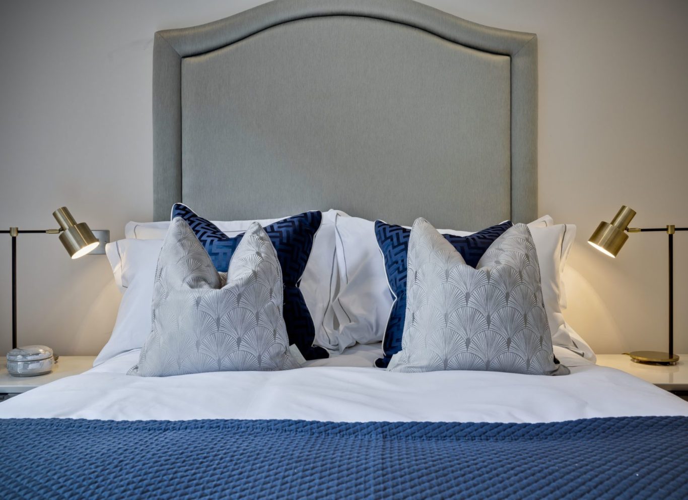 brass-bedside-lighting-luxury-headboard-surrey Symmetrical modern bedroom design by Curated Chaos Interiors featuring a curved grey headboard, navy blue textured cushions